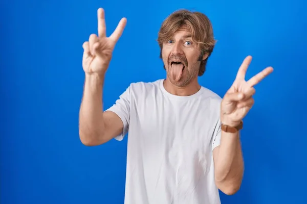 両手の指が勝利の印をしているのを示す舌で笑顔青い背景の上に立っている中年の男 — ストック写真