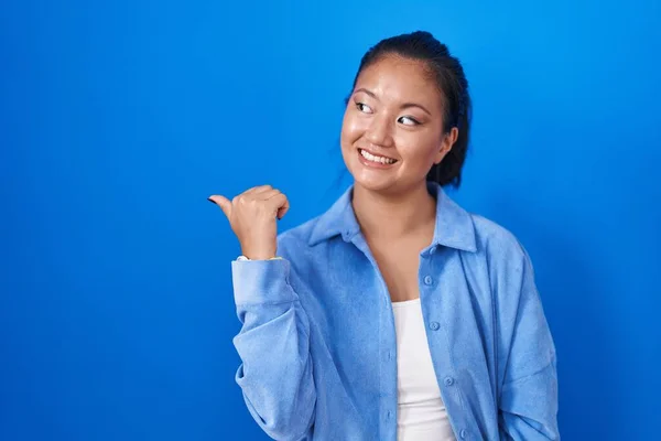 亚洲的年轻女子站在蓝色的背景上 笑容满面 高举大拇指指向一边 — 图库照片