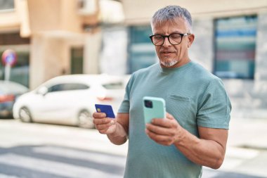 Middle age grey-haired man using smartphone and credit card at street