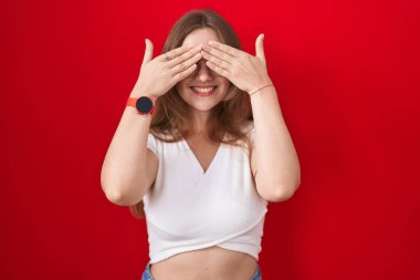 Genç, beyaz bir kadın kırmızı arka planda duruyor. Gözleri, neşeli ve komik gülümseyen ellerle örtüyor. kör kavram. 