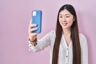 Çinli genç bir kadın akıllı telefonuyla fotoğraf çekiyor. Pozitif görünüyor ve mutlu görünüyor. Kendine güvenen bir gülümsemeyle dişlerini gösteriyor. 