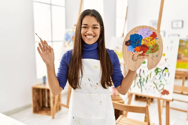 年轻的拉丁女子微笑着 自信地在艺术工作室拿着画笔和调色板 — 图库照片