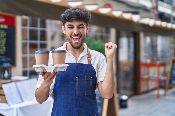 Arab man with beard wearing waiter apron at restaurant terrace screaming proud, celebrating victory and success very excited with raised arms 