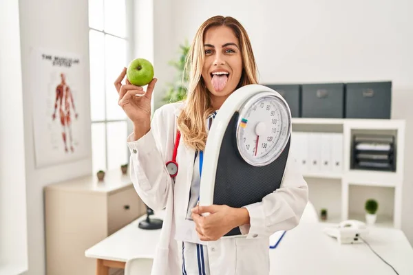 若いですブロンド医師女性保持計量機と緑のリンゴステッキング舌アウト幸せとともに面白い式 — ストック写真