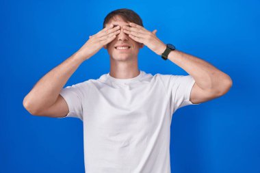 Beyaz, sarışın bir adam mavi arka planda duruyor. Gözleri kaplıyor, neşeli ve komik gülümsüyor. kör kavram. 