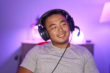 Çinli genç adam video oyunları oynuyor, kulaklık takıyor. Yan tarafa bakıyor, profil pozu veriyor, doğal bir yüzü ve kendine güvenen bir gülümsemesi var.. 