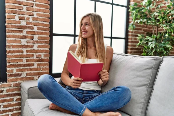 Young Blonde Woman Reading Book Sitting Sofa Home — стоковое фото