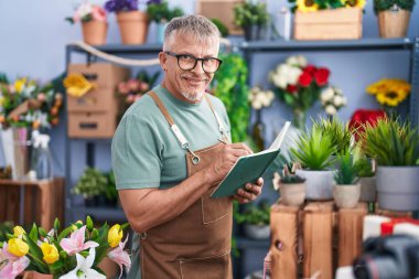 Middle age grey-haired man florist smiling confident writing on notebook at flower shop