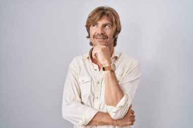 Orta yaşlı bir adam, elleri çenesinde, soyutlanmış bir arka planda durup soru sormayı, düşünceli bir ifadeyi düşünüyor. Gülümseyen ve düşünceli bir yüz. şüphe kavramı. 