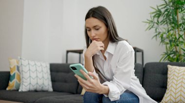 Genç, güzel, İspanyol bir kadın evde ciddi bir ifadeyle akıllı telefon kullanıyor.