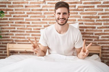 Beyaz bir adam zafer jesti yapıyor. Yatak odasında oturuyor.