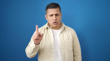 Sakin bir ifadeyle duran genç İspanyol adam, izole edilmiş mavi arka planda parmaklarıyla hayır diyor.