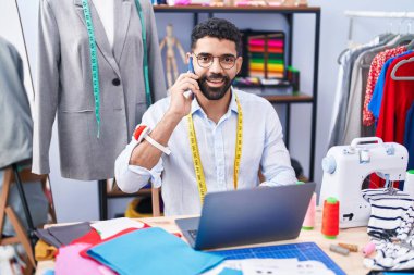 Young arab man tailor talking on smartphone using laptop at tailor shop