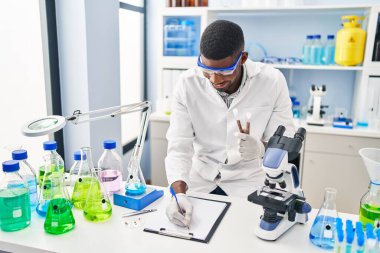 Young african american man wearing scientist uniform writing on clipboard holding test tubes at laboratory