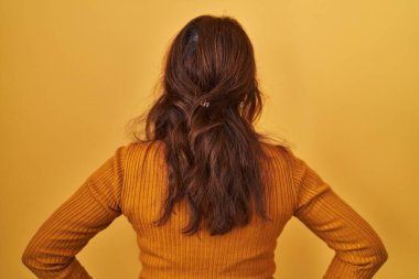 Orta yaşlı İspanyol bir kadın, sarı arka planda dikiliyor, geriye bakıyor, kolları vücudunda. 