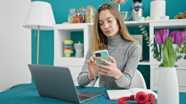 Genç beyaz kadın yemek odasında dizüstü bilgisayar ve akıllı telefon kullanıyor.