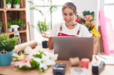 Çiçekçide çalışan genç, sarışın bir kadın. Video çekiyor. Mutluluk dolu bir gülümsemeyle başarıyı kutluyor. 