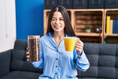 Kahve çekirdeği ve kahve fincanı tutan genç esmer kadın kameraya seksi bir ifadeyle bakıyor, neşeli ve mutlu bir yüzle.. 