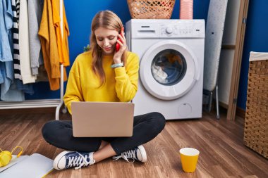 Young blonde woman using laptop talking on smartphone waiting for washing machine at laundry room