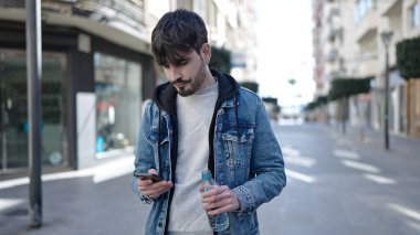 Genç İspanyol adam su içmek için caddede akıllı telefon kullanıyor.