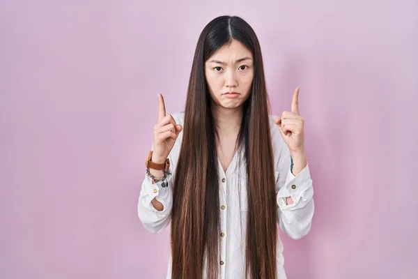 中国的年轻女子站在粉红的背景上 面带微笑地抬起头 用手指指示着方向 既不快乐又沮丧 — 图库照片