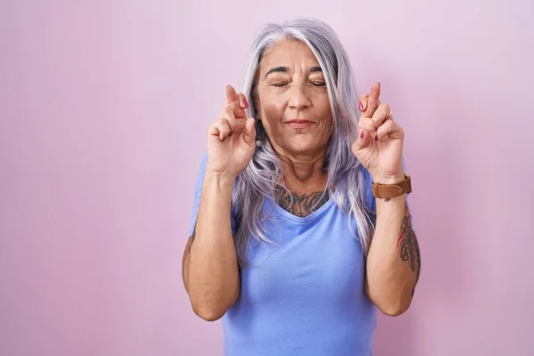 Middle age woman with tattoos standing over pink background gesturing finger crossed smiling with hope and eyes closed. luck and superstitious concept.