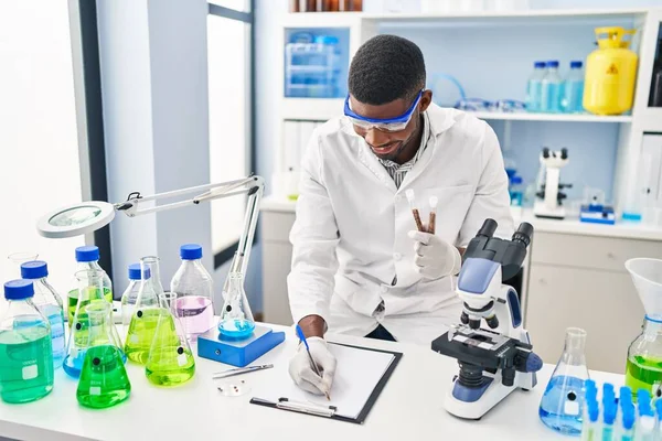 若いアフリカ系アメリカ人の男を身に着けています科学者の均一な書き込みクリップボードに保持テストチューブで実験室 — ストック写真