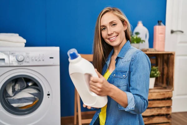 年轻的金发女人在洗衣房洗衣服 手里拿着洗涤剂瓶子 — 图库照片
