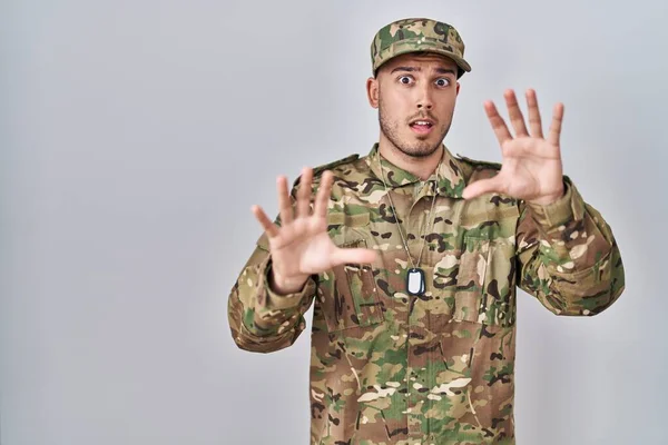 Young hispanic man wearing camouflage army uniform afraid and terrified with fear expression stop gesture with hands, shouting in shock. panic concept.