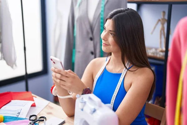 年轻美丽的惊慌失措的女人裁缝在裁缝店用智能手机自信地微笑着 — 图库照片