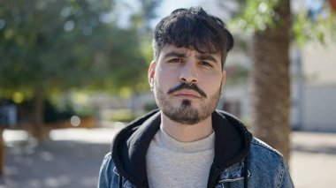 Young hispanic man standing with serious expression at park