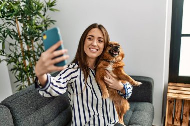 Genç İspanyol kadın akıllı telefonun başında selfie çekiyor. Kanepede oturup evdeki köpeğe sarılıyor.