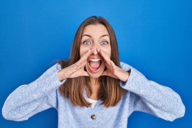 Young woman standing over blue background shouting angry out loud with hands over mouth 
