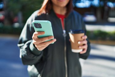 Young beautiful hispanic woman using smartphone drinking coffee at street