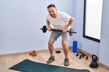 Spor merkezinde bar ve ağırlık diski kullanarak kendinden emin gülümseyen beyaz bir genç.