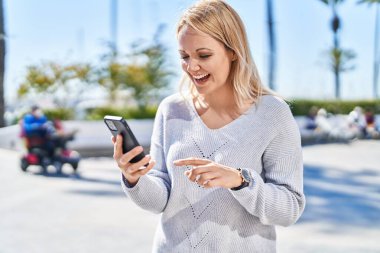 Genç sarışın kadın gülümsüyor. Parkta akıllı telefondan videolar izliyor.