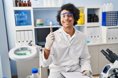 Laboratuvarda elinde pano ve test tüpü tutan bilim adamı üniforması giyen İspanyol genç adam.