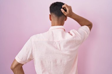 Young hispanic man standing over pink background backwards thinking about doubt with hand on head 