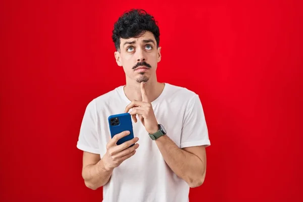 Homem Hispânico Usando Smartphone Sobre Fundo Vermelho Pensando Concentrou Sobre — Fotografia de Stock