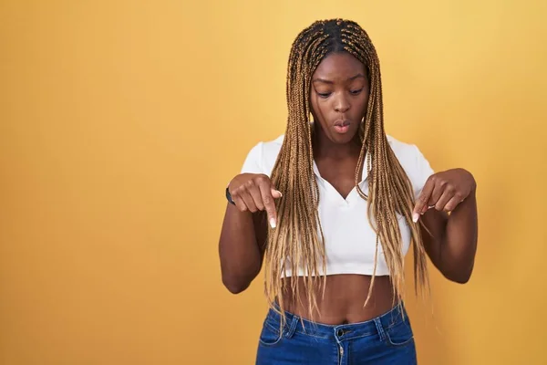 非洲裔美国妇女 辫子状的头发披在黄色的背景上 手指垂下 展示广告 惊讶的脸和张开的嘴 — 图库照片
