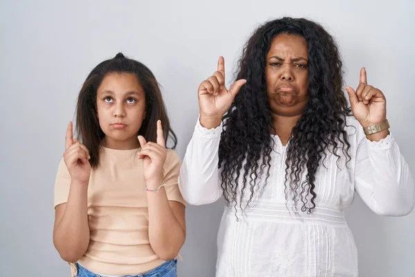 stock image Mother and young daughter standing over white background pointing up looking sad and upset, indicating direction with fingers, unhappy and depressed. 