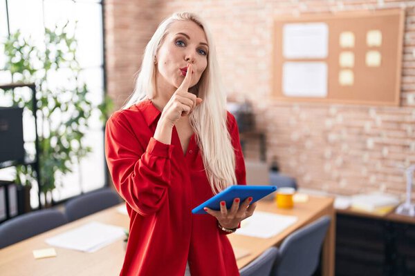 Caucasian woman working at the office with tablet asking to be quiet with finger on lips. silence and secret concept. 