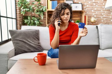 Evde kredi kartı ve dizüstü bilgisayar kullanarak akıllı telefondan konuşan genç Latin kadın.