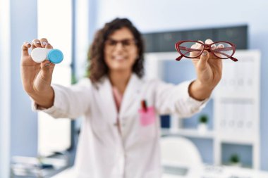 Optik klinikte gözlükçü üniforması giyen genç Latin bir kadın optik klinikte gözlük ve lens tutuyor.