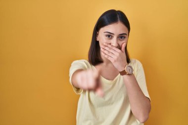 Sarı arka plan üzerine sıradan tişört giyen İspanyol kız sana gülüyor, eli ağzında kamerayı işaret ediyor, utanç ifadesi var. 