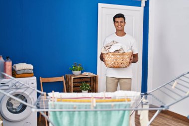 Çamaşır odasında çamaşır sepetini tutan kendine güvenen İspanyol genç adam.