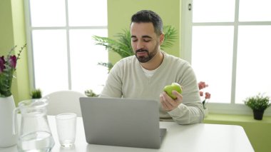 Genç İspanyol adam evde dizüstü bilgisayar kullanarak elma yiyor.