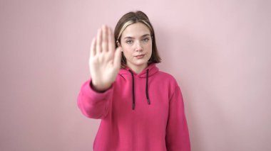 Young blonde woman doing stop gesture with hand over isolated pink background