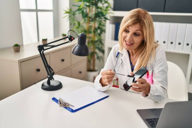 Klinikte ekranda akıllı telefon gösteren doktor üniforması giyen orta yaşlı sarışın kadın.