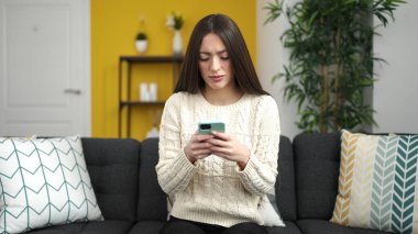 Genç, güzel, İspanyol bir kadın evde ciddi bir ifadeyle akıllı telefon kullanıyor.